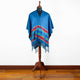 A blue poncho with colorful geometric patterns drapes over a mannequin on a wooden stand, situated in a room with light-colored walls and wooden flooring.