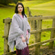 A person wearing a gray shawl stands by a rustic wooden fence in a lush green landscape, exuding a serene, contemplative mood.
