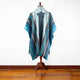 A mannequin displays a poncho with blue and gray stripes, featuring intricate patterns; it stands in a minimalist room with a wooden floor.