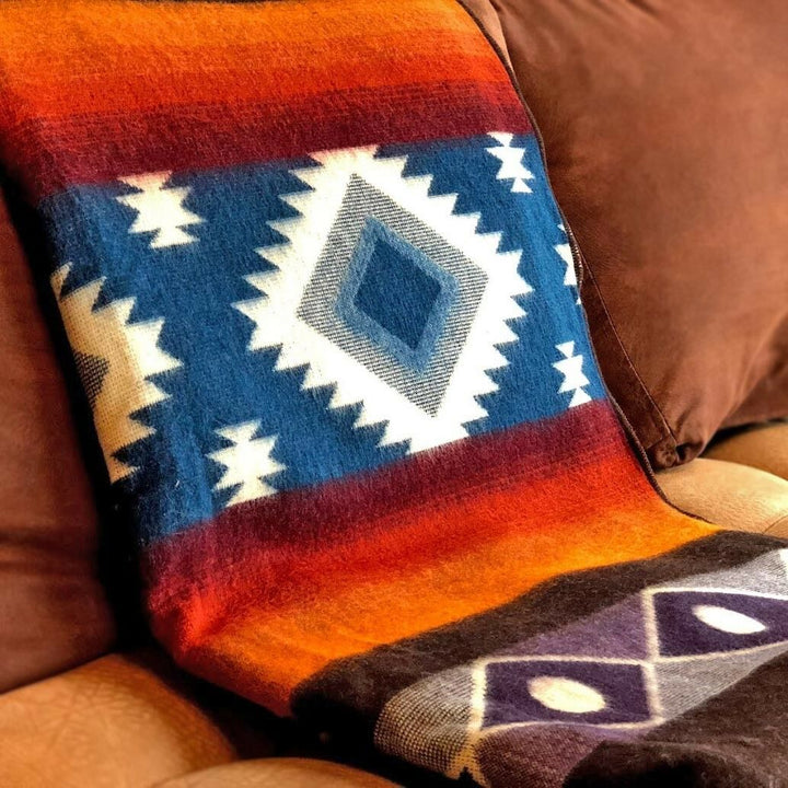Qhusi extra large alpaca wool blanket – a colorful blanket with geometric patterns is draped over a brown couch, displaying diamond shapes in blue, white, and orange hues, enhancing a cozy living room setting