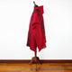A red poncho with black geometric patterns is displayed on a mannequin. It features fringe details and stands on a wooden floor against a plain white background.