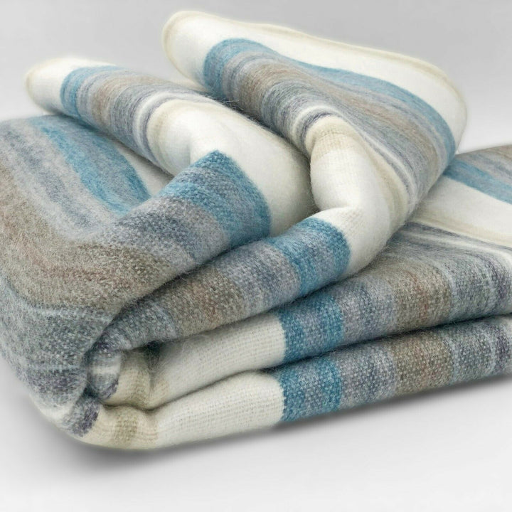 Chuquipogyo baby alpaca wool blanket – a folded woolen blanket displays horizontal stripes in shades of blue, gray, and beige, resting on a flat, neutral surface