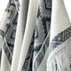 Cuyuja lightweight alpaca wool hooded poncho – a white poncho with intricate black geometric patterns and fringed edges is draped, showcasing its design and texture. It is set against a plain background