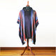 A multicolored striped poncho with a hood and button detail is displayed on a wooden mannequin in a room with a wooden floor and a plain white wall background.