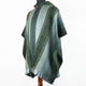 A green, striped poncho with embroidered patterns drapes over a mannequin in a minimalist setting. The garment features a hood and a button closure at the neck.