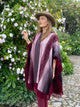 A person stands amidst blooming flowers, wearing a wide-brimmed hat and a patterned poncho. They gently touch a flower, surrounded by lush greenery and a cobblestone path.