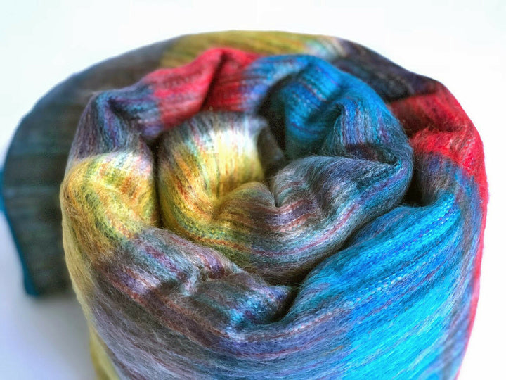 Alpaca wool queen blanket – a rolled-up, multi-colored woolen fabric sits on a plain white surface, showcasing vibrant hues of red, blue, yellow, and green intertwined in a soft, textured spiral