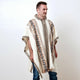 A person is wearing a patterned poncho with geometric designs, standing against a plain white background. The poncho features an intricate design and fringe at the bottom.