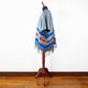 A colorful poncho with geometric patterns and fringe detail is draped over a mannequin. It stands on a wooden floor against a plain white background.