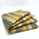 Rumi baby alpaca wool blanket – a folded multicolored striped blanket lies neatly stacked against a plain white background, showcasing an array of muted greens, yellows, and oranges