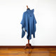 A blue poncho with a hood is displayed on a wooden mannequin stand against a plain white wall. The polished wooden floor adds warmth to the minimalistic setting.