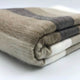 Mogato baby alpaca wool blanket – a folded, woolen blanket featuring stripes in shades of beige, gray, and white, resting on a flat, plain surface