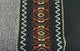 Llama Wool Hooded Poncho – textile with intricate geometric patterns, featuring orange, beige, and black motifs. Patterned stripe flanked by zigzag designs on dark green and gray textured background