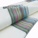 Sigchos baby alpaca wool blanket – a folded white fabric is displayed, featuring horizontal stripes in various colors like green, pink, and blue. The fabric is positioned on a plain surface, filling the frame