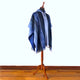 A blue poncho with intricate patterns drapes over a mannequin, standing on a wooden floor against a plain, light-colored background.