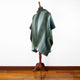 A green poncho with striped patterns drapes on a mannequin. Wooden floor and white wall provide a minimalist background.