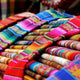 Stacks of vibrant, multicolored woven fabrics are layered neatly, showcasing intricate patterns. The textiles create a visually striking and colorful display, likely in a market setting.