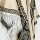 A fringed textile features geometric patterns in gray and black tones, hanging against a white background. The fabric showcases diamond shapes in a soft, woven material.