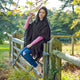 A person sits on a wooden fence, holding a branch, wearing a shawl and jeans. The background features a lush green landscape with trees and scattered sunlight.