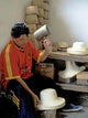 A person in a red shirt shapes a hat with a wooden mallet, seated beside a table holding several hats and boxes. Hats are stacked in the background.
