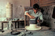 A man is shaping a straw hat with an iron, focusing intently at a wooden workbench in a rustic workshop, surrounded by stacked hats and fabric.