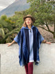 A person in a blue poncho and hat stands against a white wall. They are outdoors, surrounded by trees, with a mountain in the background.