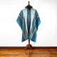 A colorful poncho with geometric patterns is displayed on a wooden mannequin stand, positioned on a polished wooden floor against a plain white background.