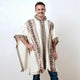 Man wearing a patterned poncho smiles with one hand on hip, standing against a plain background. The poncho is cream with geometric designs in earthy tones.
