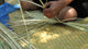 Hands weave thin palm leaves on a woven mat, with a blue sheet partially visible, suggesting outdoor crafting activities.