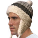 A man wears a knitted winter hat with ear flaps and a pom-pom, featuring a beige fluffy lining and dark patterned exterior, against a plain white background.