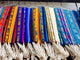 Colorful woven textiles hang on hangers, showcasing intricate patterns and vibrant stripes in blues, yellows, and reds, with white fringes at the bottom. They are displayed in a market setting.