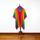 A colorful poncho with wide rainbow stripes hangs on a mannequin stand, set against a plain white wall and wooden floor.