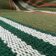 A woven textile with green, white, and orange stripes lies flat, showing detailed texture and pattern. The background consists of darker and lighter bands.