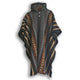 A knitted hooded poncho features geometric patterns in orange, brown, and black with white zigzag lines. It hangs on a black hanger, displaying its intricate woven design in a minimalist setting.