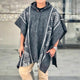 A person wears a detailed black and white patterned poncho while standing on a tiled surface indoors, with a blurred cityscape visible in the background.