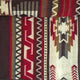 A woven textile displaying intricate geometric patterns in red, brown, and white hues. The design includes zigzags, chevrons, and abstract motifs, creating a visually dynamic and textured appearance.
