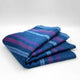 Tamboloma baby alpaca wool blanket – folded woolen blanket featuring blue, purple, and teal stripes, resting on a smooth, white surface. The blanket appears soft and plush, suggesting warmth and comfort
