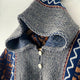 A knitted sweater with a hood hangs on a light background. It features blue and gray yarn, white buttons, and intricate orange and white geometric patterns on the sleeves.