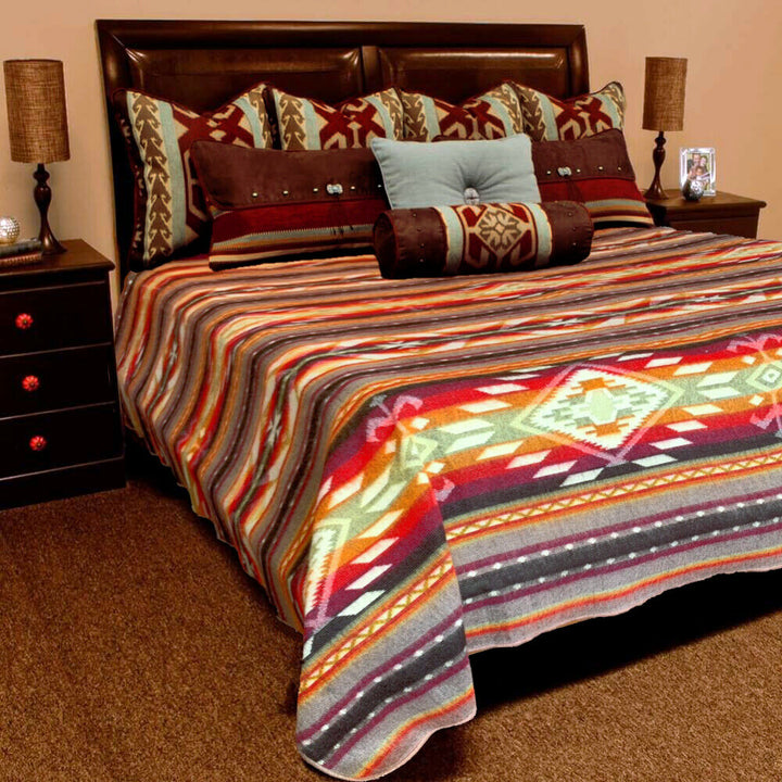 A bed displays a colorful, geometric-patterned blanket, surrounded by multiple decorative pillows, in a cozy bedroom with brown nightstands, each holding a lamp and small decorative items.