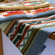 A multicolored patterned blanket with geometric designs drapes over a wooden table, contrasting its vibrant hues against the neutral indoor setting.