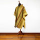 A yellow and orange patterned poncho drapes over a mannequin stand, displayed in a room with wooden flooring and a plain white wall background.