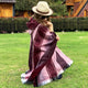 A person wearing a hat and sunglasses wraps a striped, maroon poncho around their shoulders on a grassy lawn, with a wooden fence and house in the background.