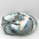 Chuquipogyo baby alpaca wool blanket – a soft, striped wool blanket is loosely coiled, showcasing shades of blue, gray, and cream. It rests against a plain white background, creating a calm, cozy atmosphere