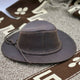 A brown leather hat with a braided band rests on a textured fabric featuring white and brown patterns, including geometric shapes and letters.