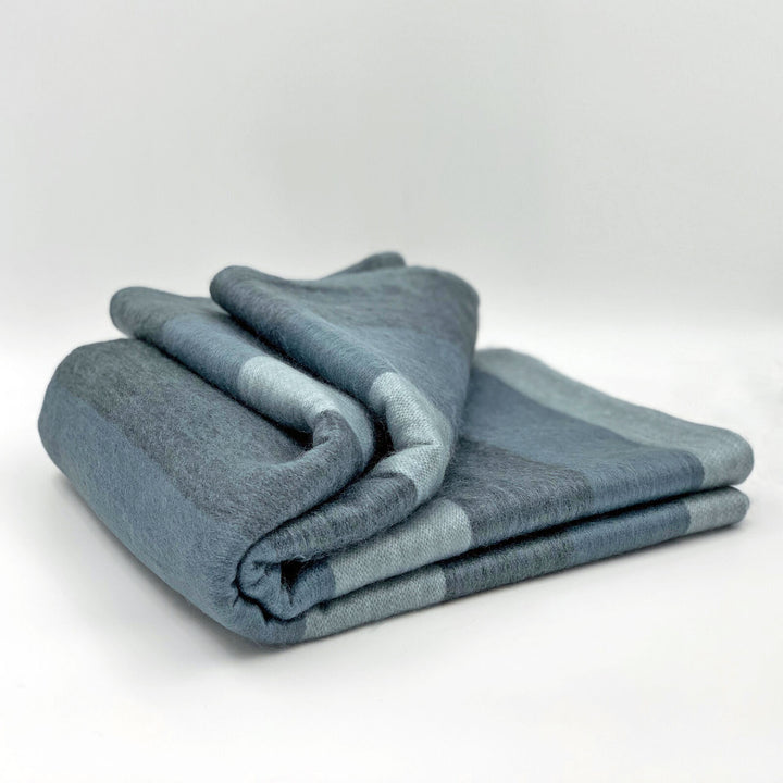 Toacazo baby alpaca wool blanket – a folded, blue-gray wool blanket rests on a plain white surface, showcasing its soft texture and subtle striped pattern