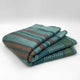 Pallalanga baby alpaca wool blanket – folded wool blanket featuring teal, brown, and green horizontal stripes resting on a smooth, light gray surface, creating a soft, inviting appearance