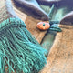 A close-up of a woolen coat with a large wooden button. It features green yarn fringes hanging over the buttoned area, creating a cozy, textured appearance.
