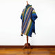 A colorful poncho with geometric patterns drapes over a wooden mannequin stand, set against a plain white wall and wooden floor, highlighting its vibrant design.