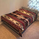 A metal-framed bed is covered with a geometric-patterned, multicolored blanket. It is situated in a carpeted room with floral curtains on the window nearby.
