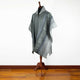 A grey poncho with geometric patterns is displayed on a wooden mannequin stand in a room with a wooden floor and white walls.
